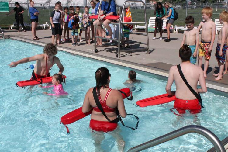 Photo shows lifeguards helping children in pool while other kids wait on pool deck at Water Safety Day.