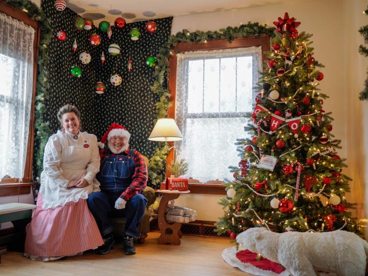santavillage6 Santa and Mrs. Claus sitting in house on Santa's chair next to lamp and Christmas Tree decorated