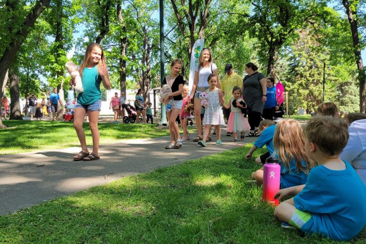 Kids and adults walking through Island Park with their stuffed animals for the Teddy Bear Parade while other families sit on the grass and watch