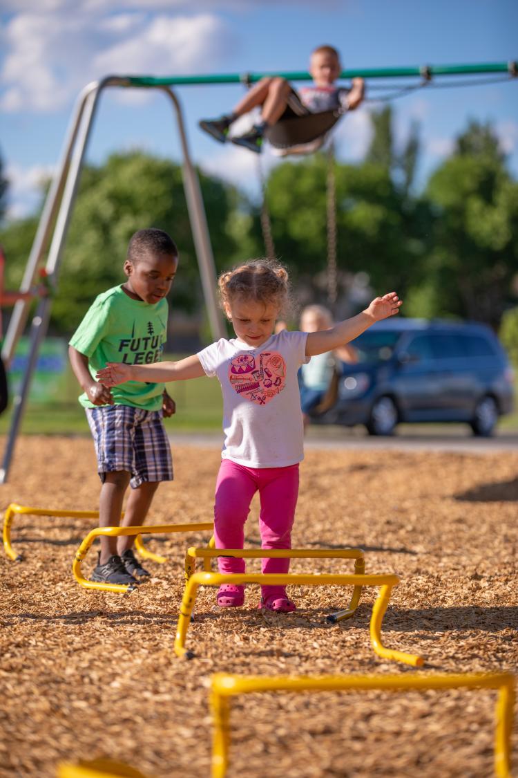 Little girl and little boy walking over little hurdles in park with boy swinging in background