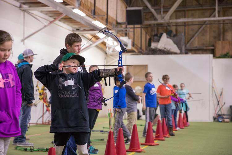 Young boy pulling back bow to shoot arrow with instructor standing behind and many kids lined up to shoot their arrow