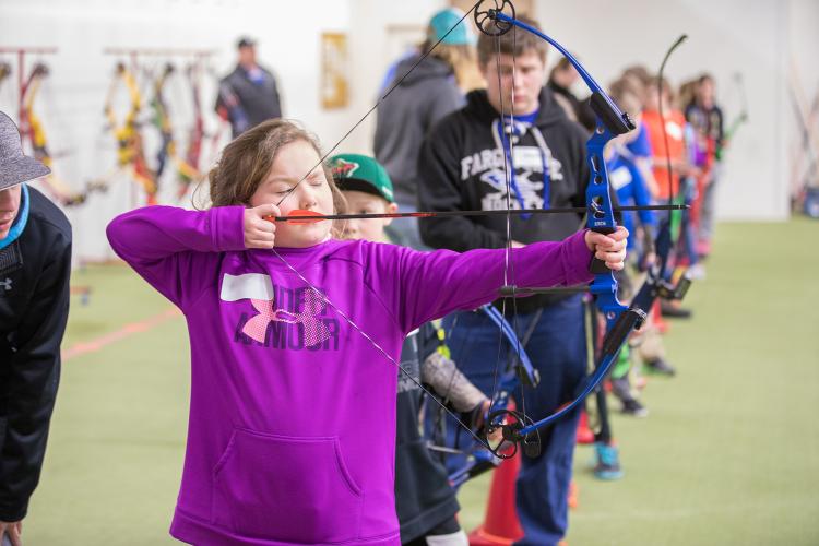 Young girl lining up bow and arrow to shoot with many other kids in the background lined up working on getting their bow and arrow ready to shoot indoors at archery lessons