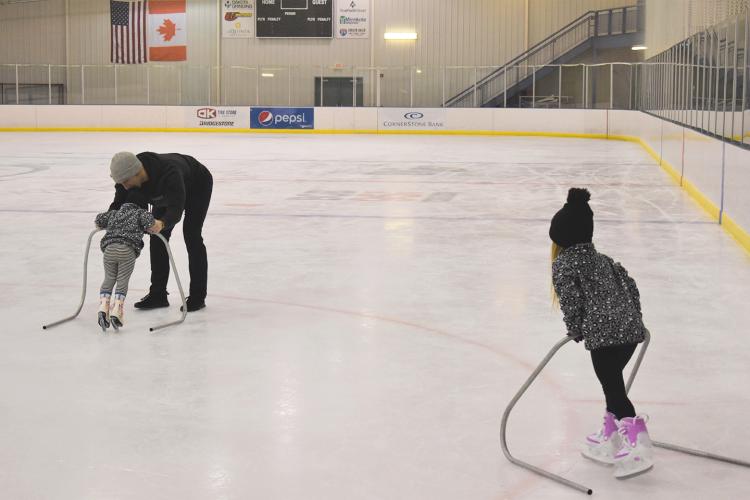 OpenSkate8.jpg two young kids ice skating indoors at the Cornerstone Bank Arena using metal skate trainers with a male adult assisting one child skate