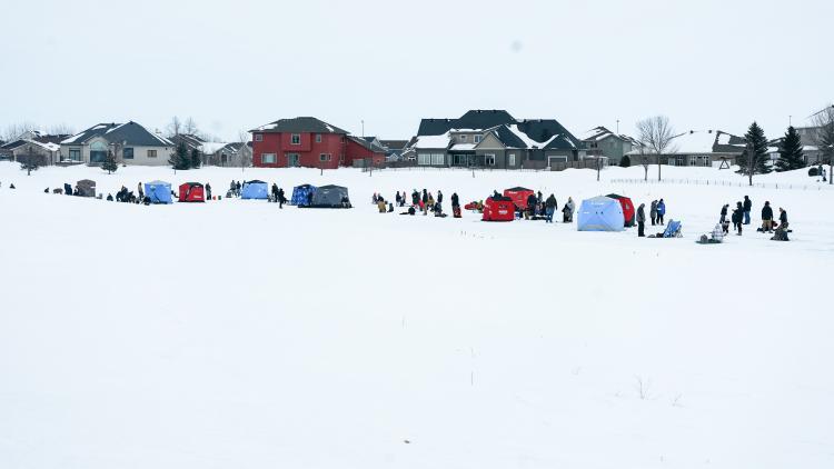 Youth Ice Fishing Derby 1 10+ ice fishing houses on the Woodhaven South Pond with 30+ people, including kids standing around on the frozen pond.  5+ large houses in the background facing the frozen pond.