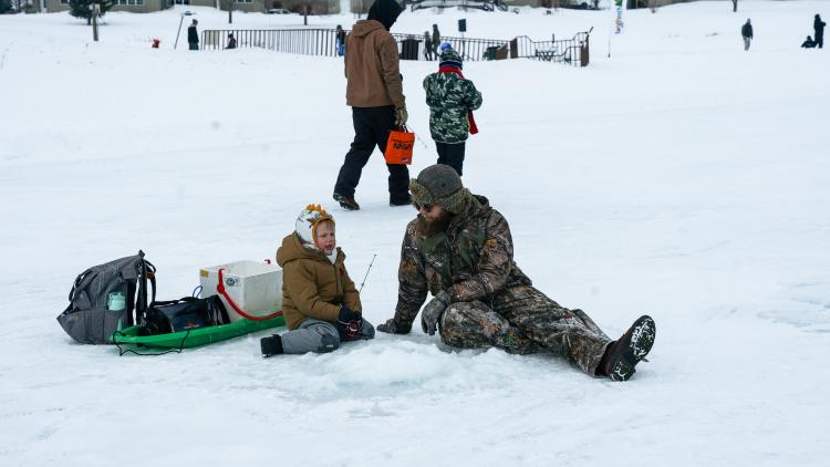 Youth Ice Fishing Derby 2 A boy and man sitting on the frozen pond next to a drilled ice fishing hole talking. The boy has a fishing pole in his hand and a sled full of ice fishing equipment sits behind the boy. In the background a boy and adult man walk .