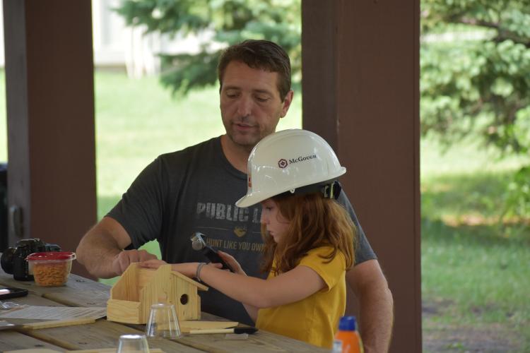 A Father and Daughter sitting at a picnic table putting together a wooden birdhouse.  The daughter is in a hard hat and yellow shirt.