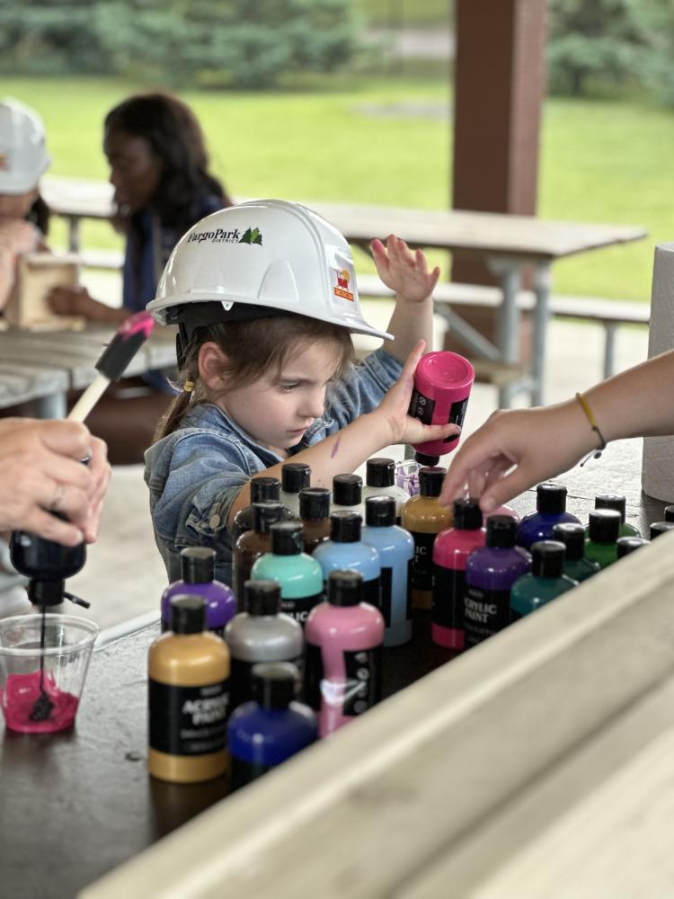 Young girl picks out the perfect paint color at a counter with 20+ paint bottles for her wood bird house, wearing a construction hard hat.