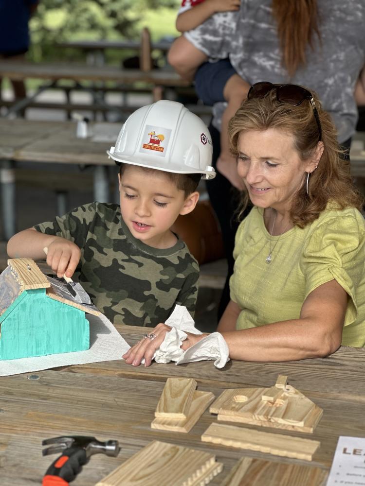 A woman sits with a young boy at a picnic table while they paint their bird house.  The boy is wearing a hard hat.