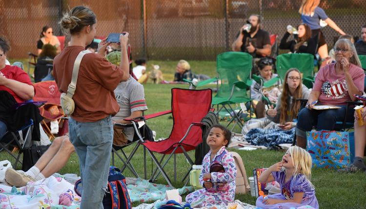 A woman stands and takes a picture of the two young girls sitting on a blanket in their PJs waiting on the lawn at Rheault Farm for the Movie in the Park movie to start with 20+ people in the background