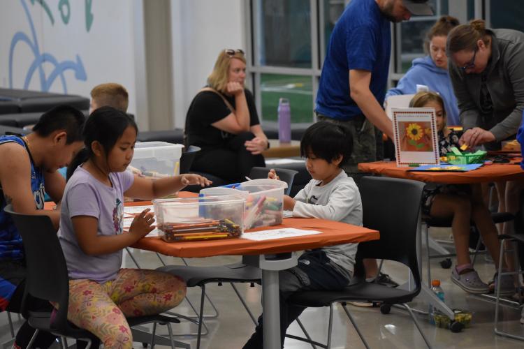 A young boy and a young girl sitting at a table with an orange table cloth doing crafts in a room full of people.