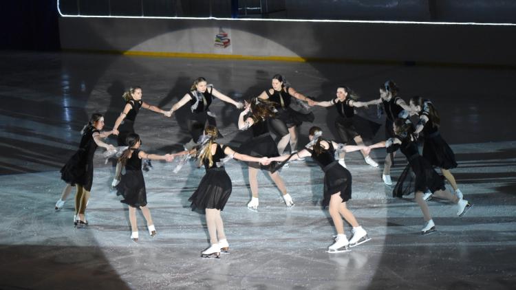11 older teens skating in a circle in black dresses around a girl in the middle skating in a spin, ice skating to a routine for the Fargo Parks Ice Revue