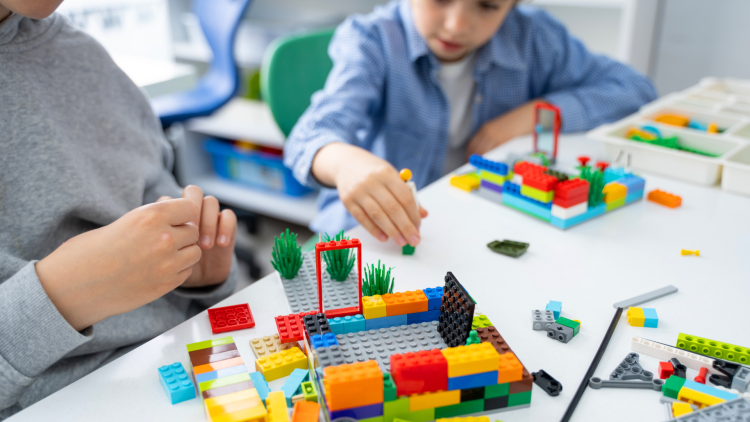 Two kids sitting in chairs at a white table building with legos.