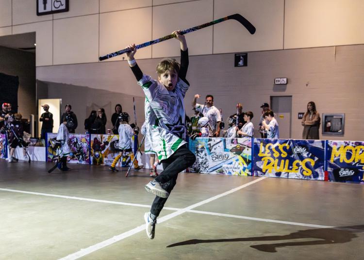 Child in jersey jumping in the air with a hockey stick over his head with parents and kids standing behind the boards cheering kids playing NHL Street on