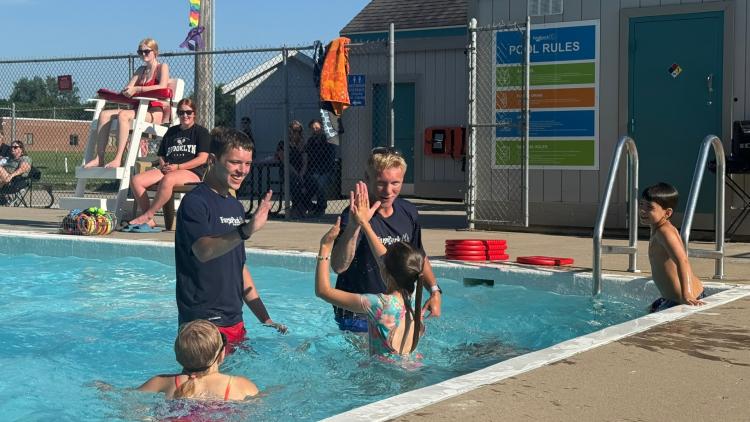 Two male lifeguards in the Madison Pool with three kids taking swimming lessons. The two guys are giving the kids high fives in the water