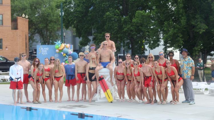 Large Group of Men and Women Lifeguards standing in a group around a few men lifeguards standing on a white tall lifeguard chair in front of the new Island Park Pool