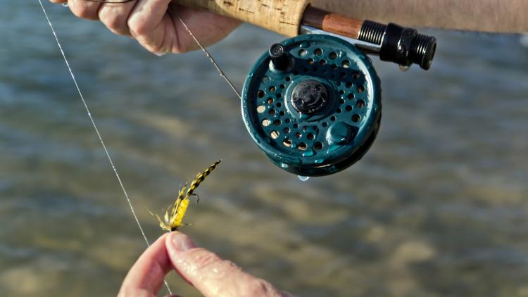 Close-up of hands holding a fly fishing rod and reel while tying a yellow-and-black fly lure, with water visible below.