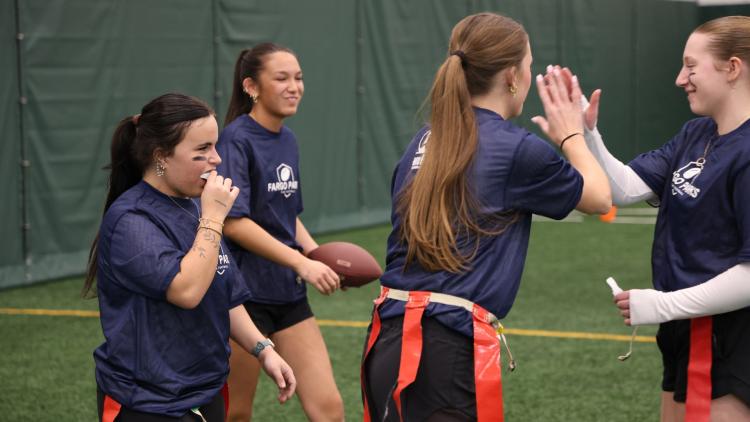 Four high school girls in practice shirts and flag belts smile and high-five during a flag football practice indoors.