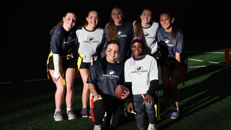 Group of high school girls wearing Fargo Parks flag football shirts pose together on an indoor turf field, smiling and holding a football.