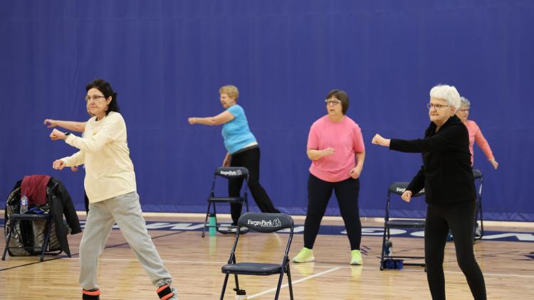 Group of older adults doing balance exercises in a gym with chairs to help maintain balance.