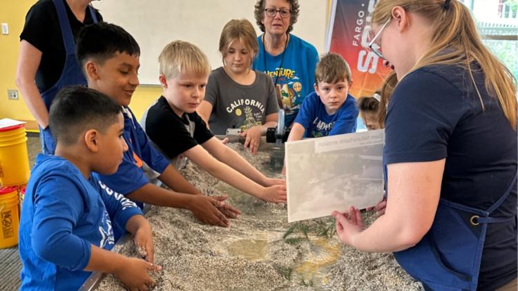 A group of kids standing around a mock river bed with sand and water running through learning from a staff member about dam erosion.