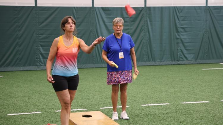 Two women play bean bag toss indoors on a turf field during the North Dakota Senior Games.