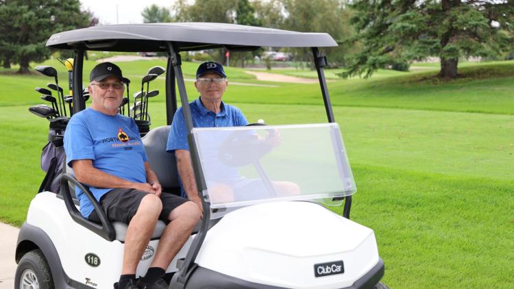 This image shows two older men siting in a golf cart on a golf course during the North Dakota Senior Games.