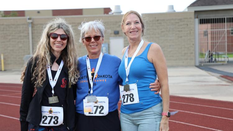 Three women wearing medals smile together on a track after competing in the North Dakota Senior Games.