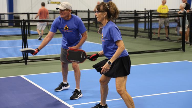This image shows two senior athletes playing pickleball on an indoor court during the North Dakota Senior Games.