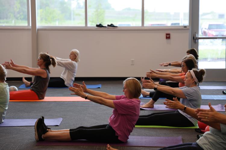 Adults participate in a group fitness class, seated on individual exercise mats with their legs extended while reaching their arms forward during a stretching and core exercise.