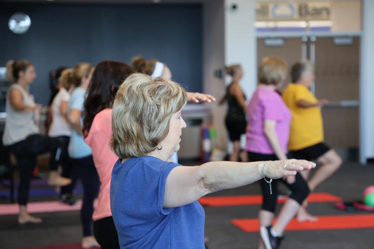 Adults participate in a group balance and fitness class, standing on individual mats while extending their arms and practicing balance poses.