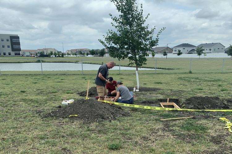 Three people work together on a landscaping project near a young tree, installing a wooden border around a planting area. A pile of soil, tools and caution tape surround the work area.