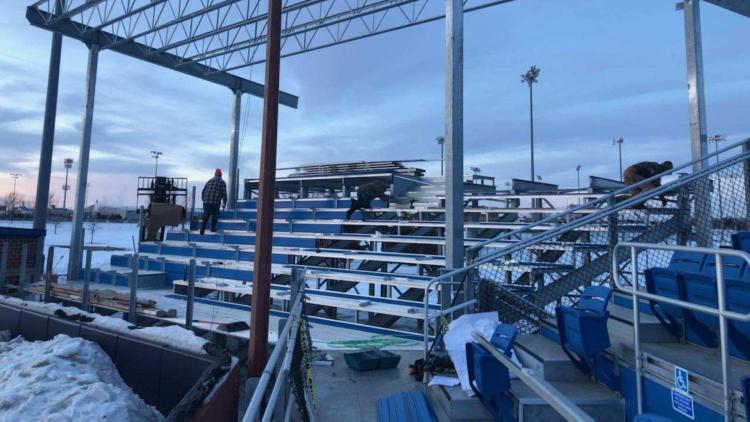 Workers install and repair aluminum bleachers at an outdoor sports complex during winter, with snow on the ground and stadium light poles in the background.