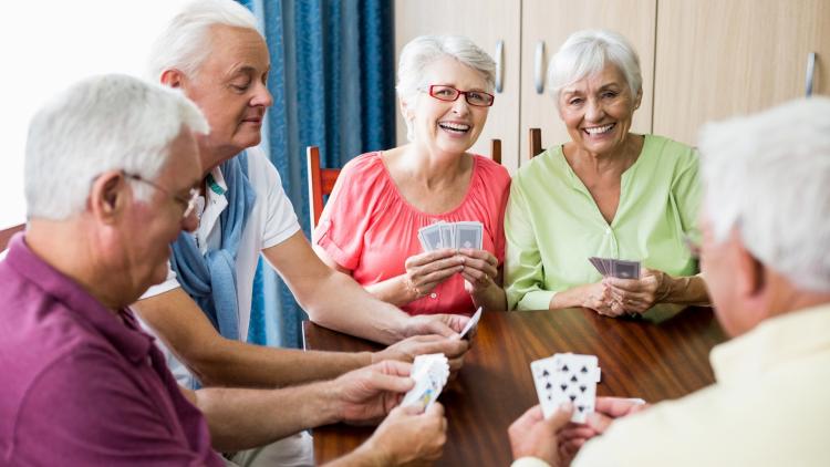 A group of older adults gathered around a table playing cards together. Several participants hold cards while smiling and socializing.
