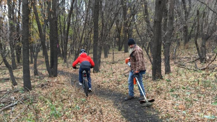 A cyclist rides along a narrow wooded trail while a park worker uses a string trimmer to maintain the trail. Fallen leaves cover the surrounding forest floor.
