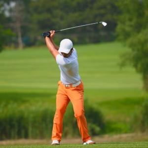 A young man golfing on a green course wearing orange pants and a white shirt and hat