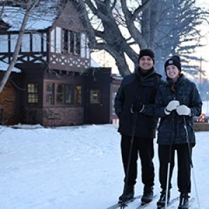 Two people smile while standing on cross-country skis along a snowy park trail at dusk, with a dog walker in the background.
