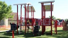 Playground at Rheault Farm Park with a farm-themed design, featuring bright red structures topped with white roofs. Panels display images of a horse and a chicken, and a slide and climbing elements are visible. The playground sits on green turf with farm buildings and trees in the background on a sunny day.