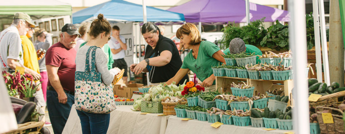 Photo shows two women assisting a woman shopping a vegetable booth at Red River Market. 