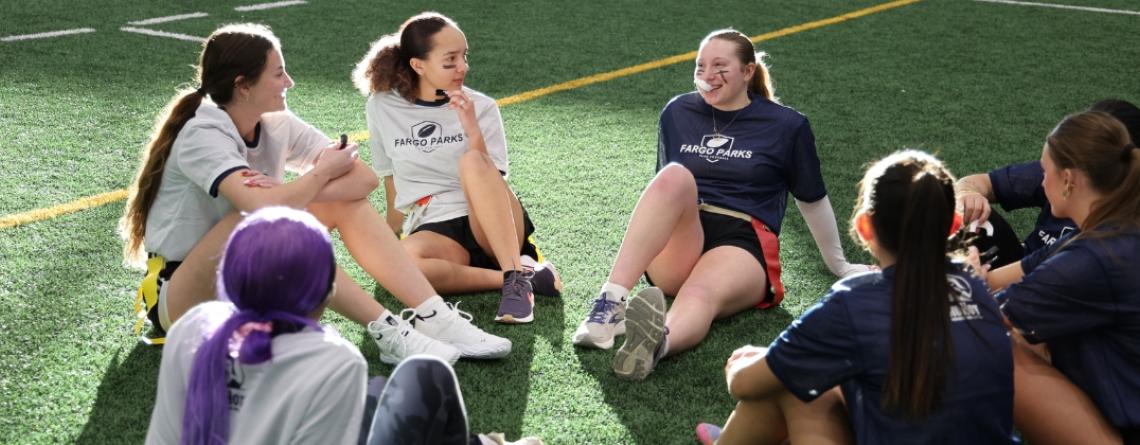 Group of high school girls in Fargo Parks flag football shirts sit in a circle on a turf field talking and relaxing during practice.
