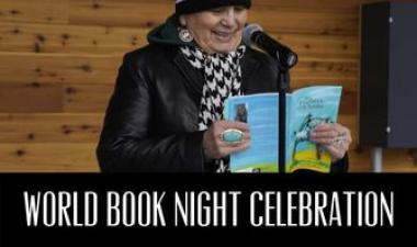 This image shows a woman reading her book into a microphone with a black bar below her with text reading World Book Night Celebration.