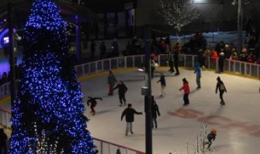 People of all ages skate on an outdoor ice rink at night at Broadway Square, surrounded by spectators and holiday lights, with a large blue-lit Christmas tree in the foreground.