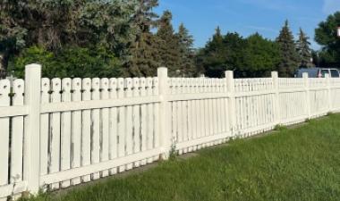 A long white picket fence stretches across a grassy area at Yunker Farm Park, with mature trees, a blue sky and a parked vehicle in the background.