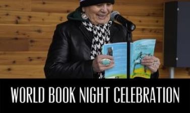 This image shows a woman reading her book into a microphone with a black bar below her with text reading World Book Night Celebration.