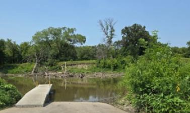 A concrete boat ramp extends into a calm river, surrounded by green trees and shrubs on a sunny summer day. Driftwood is visible along the opposite shoreline.