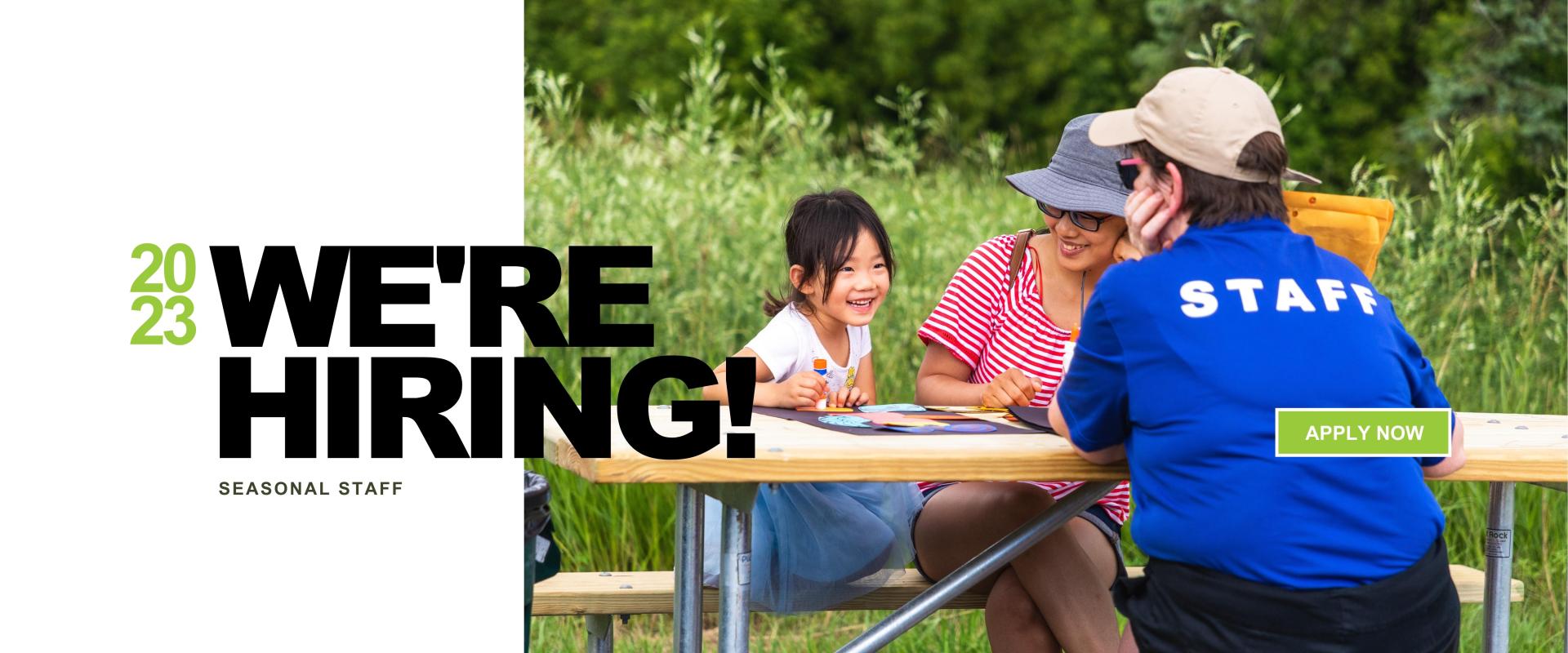 2023 WE'RE HIRING - SEASONAL STAFF - over a picture of a staff member in a blue shirt sitting at a table with a woman and a child coloring in a park