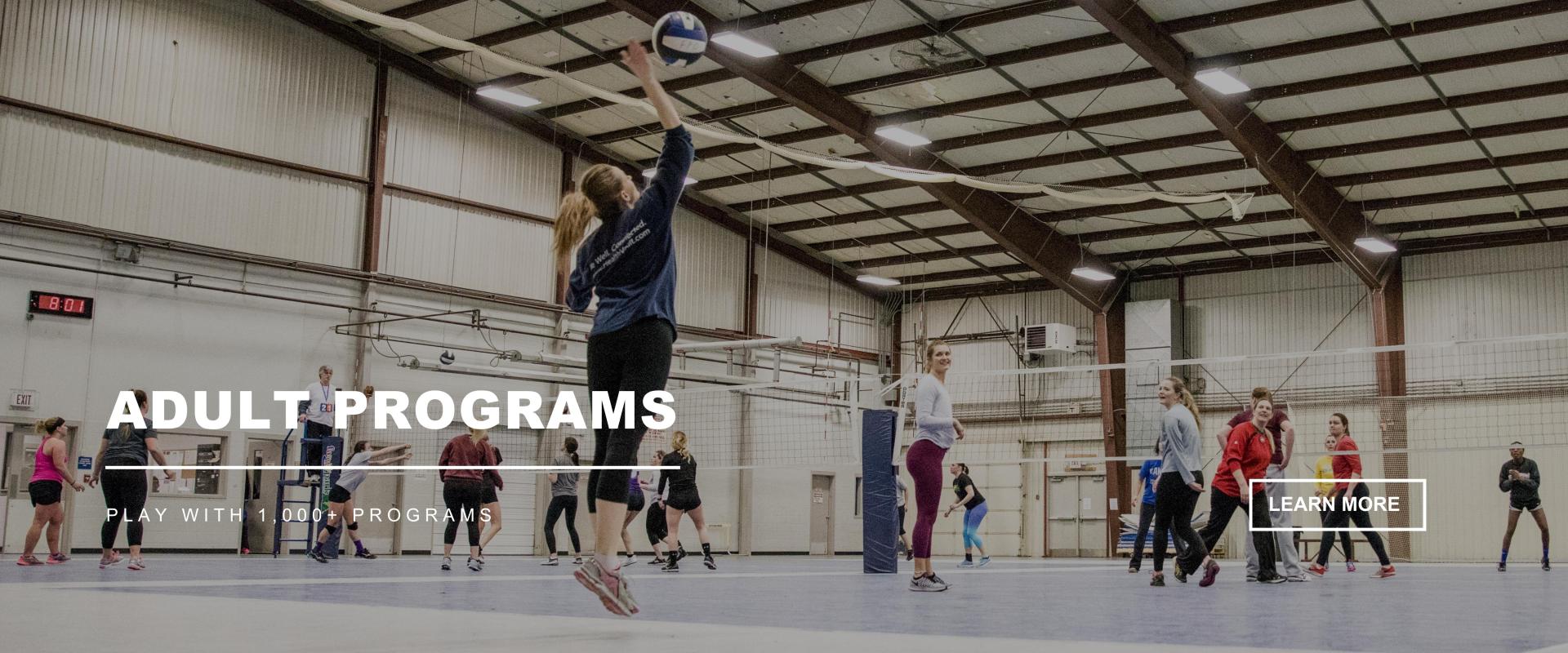 A female serving a volleyball to a team in a large building - ADULT PROGRAMS - LEARN MORE