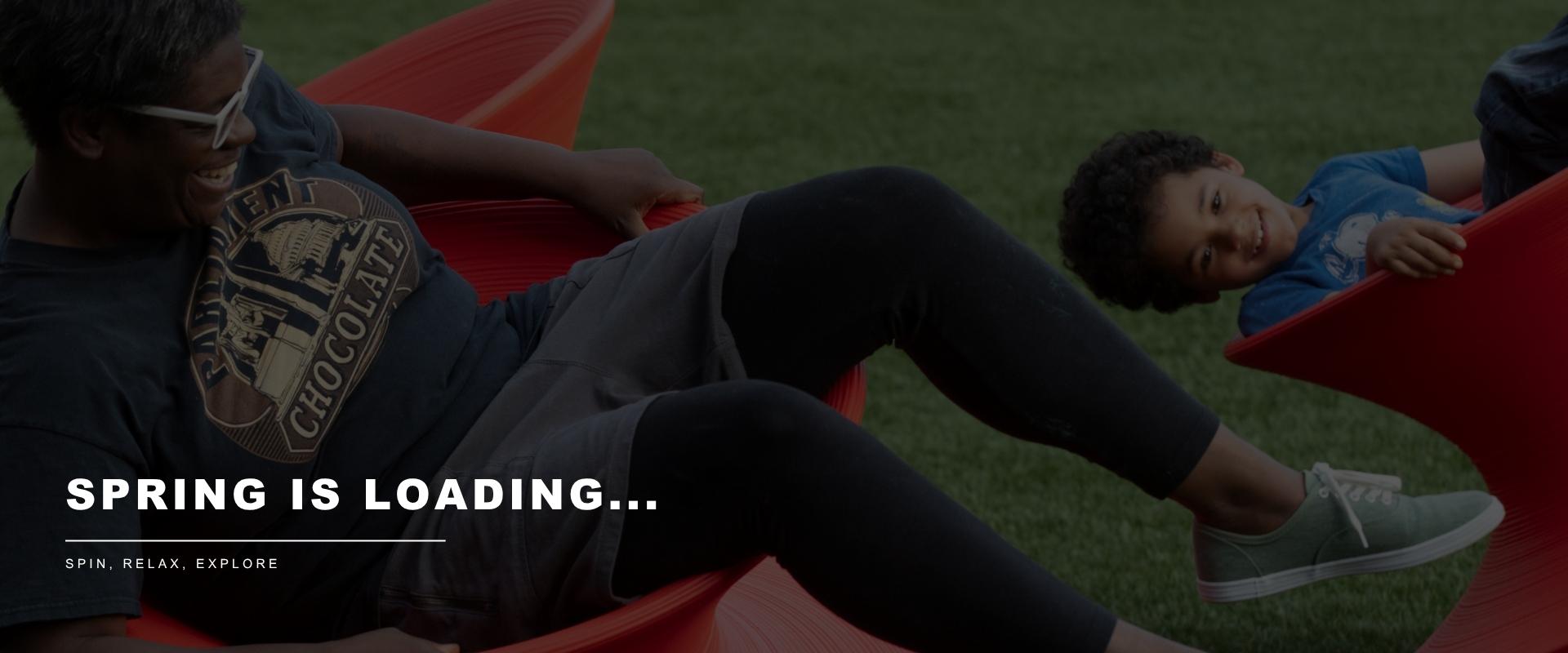 Adult and child smiling while spinning on red playground equipment outdoors. Text reads “Spring is Loading…” and “Spin, Relax, Explore.”