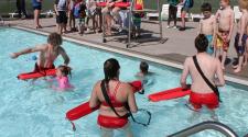 Photo shows lifeguards helping children in pool while other kids wait on pool deck at Water Safety Day.