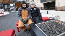 This image shows a male and female sitting on a bench by a firepit at Broadway Square.