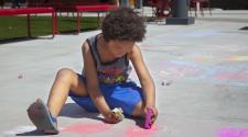 This image shows a young boy drawing on the cement with a piece of sidewalk chalk in both hands.